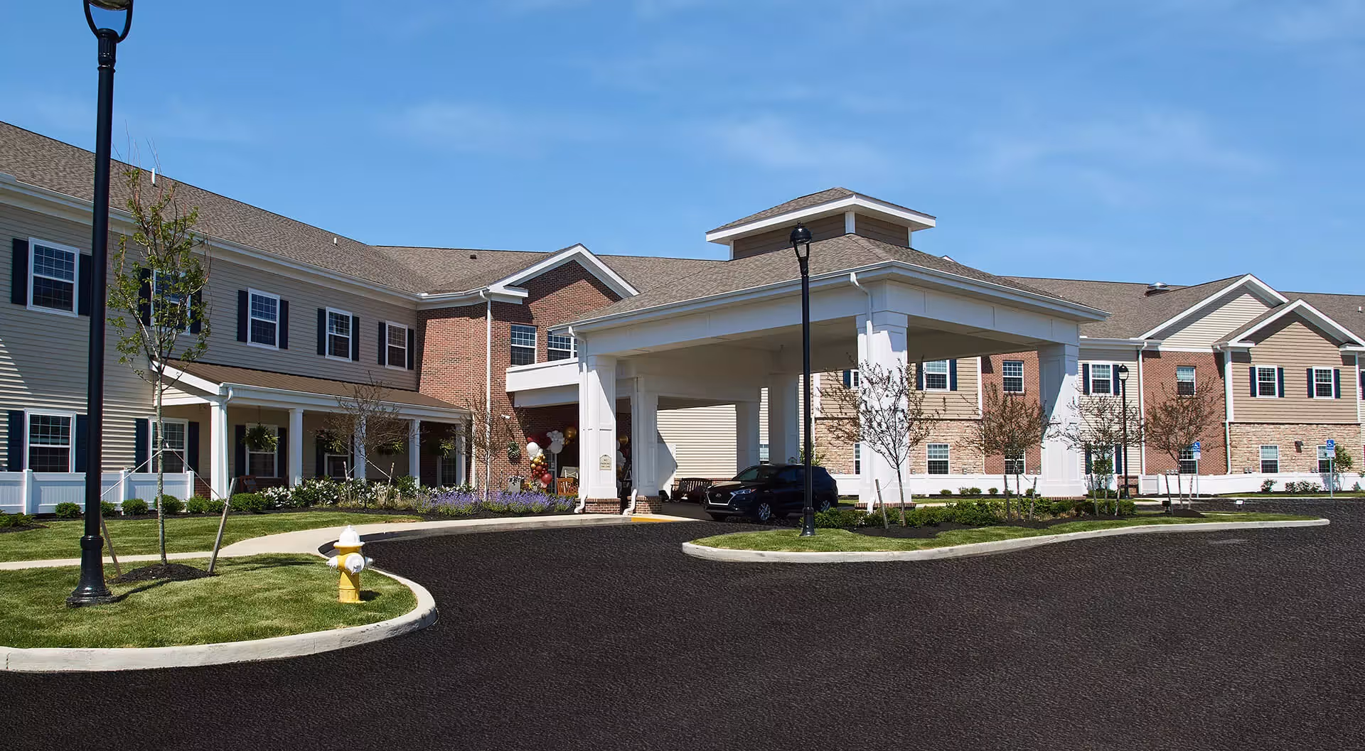 Exterior view of a senior living facility building with a covered entrance, multiple windows, and a well-maintained lawn with small trees and a fire hydrant. A black vehicle is parked under the covered entrance.