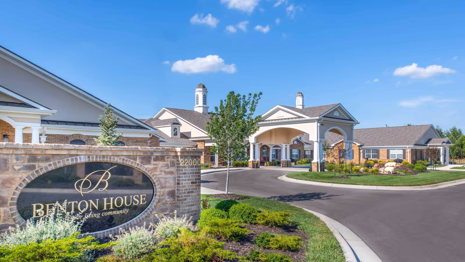 Exterior view of Benton House senior living community showing a brick sign with the community name and a driveway leading to a large building with columns and cupolas under a blue sky.