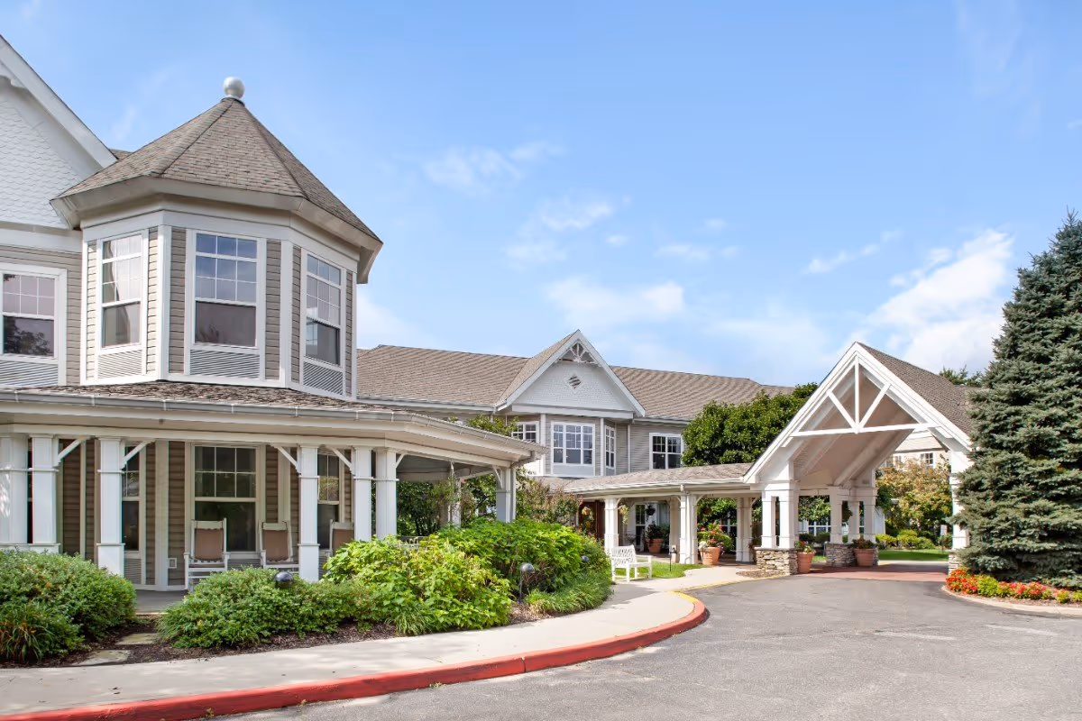 Exterior view of a senior living facility with a covered entrance, large windows, and a landscaped garden area with bushes and a tall evergreen tree under a partly cloudy sky.