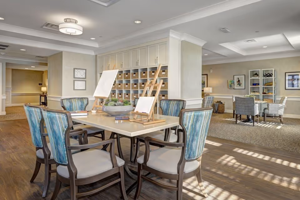 A bright and spacious common area in a senior living facility with a large table surrounded by six chairs with blue and green striped upholstery. On the table are two easels with blank canvases and a decorative bowl with succulents. In the background, there are shelves with baskets and another seating area with a table and chairs, artwork on the walls, and soft lighting from lamps.