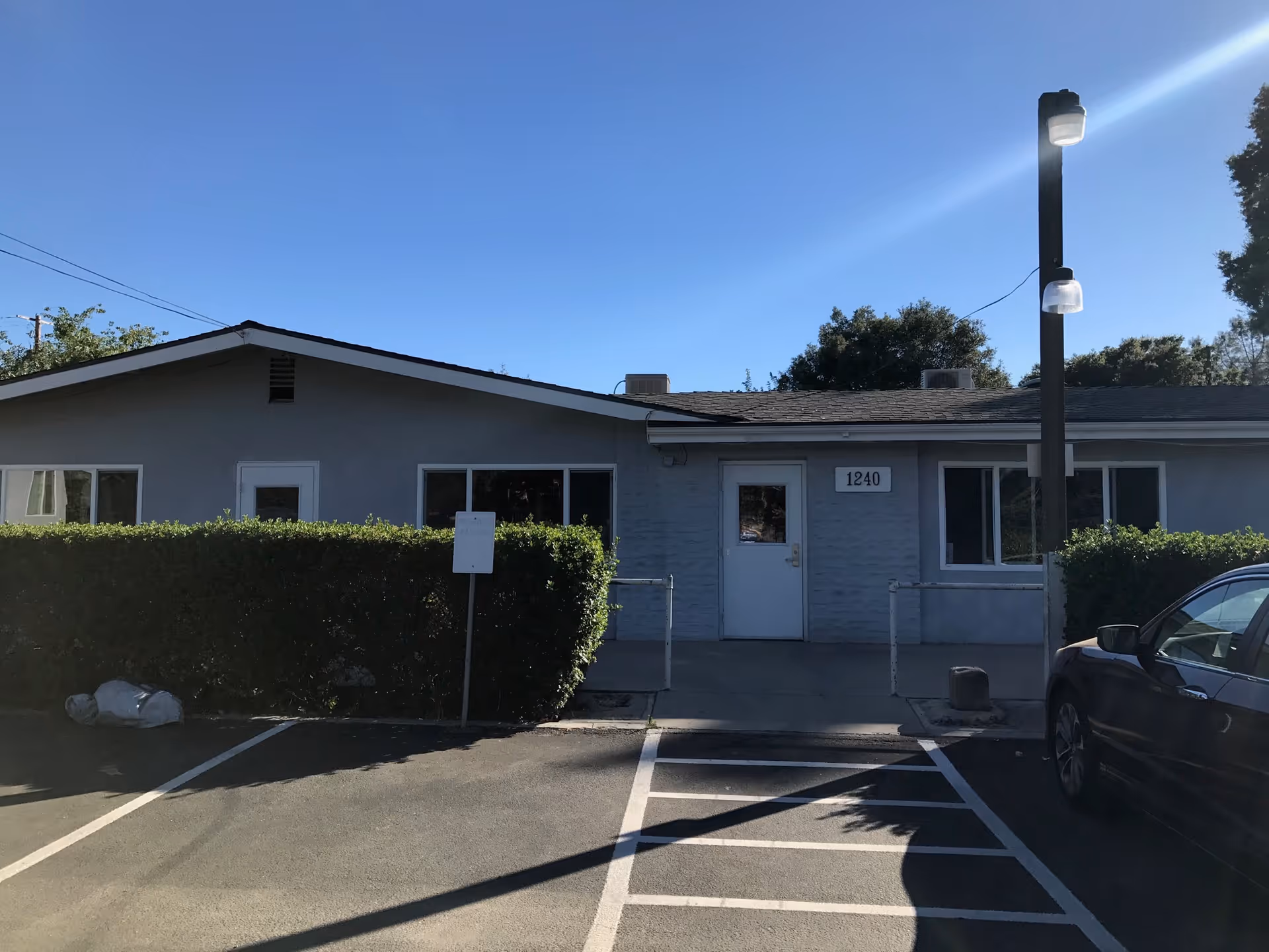 Exterior view of a single-story building with a gray facade, white door, and windows. The building has the number 1240 displayed above the door. There is a parking lot in front with marked parking spaces, a hedge along the front of the building, and a tall street lamp on the right side. A car is partially visible on the right side of the image. The sky is clear and blue.
