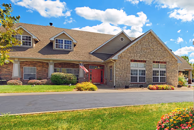 Exterior view of a single-story senior living facility building with a combination of brick and stone facade, multiple windows, a red front door, an American flag near the entrance, and a well-maintained lawn with flowers and shrubs under a blue sky with scattered clouds.