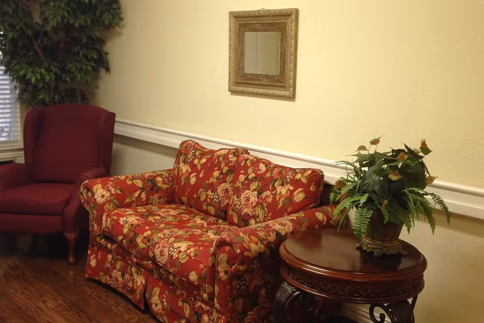 A cozy sitting area with a floral patterned red loveseat, a matching red armchair, a round wooden side table with a potted green plant, a framed mirror on the wall, and a large leafy plant in the corner near a window with blinds.