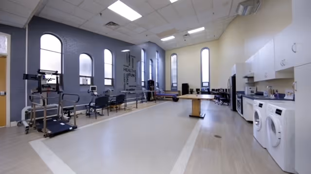 A spacious rehabilitation or therapy room with exercise equipment including a treadmill, parallel bars for walking practice, chairs along the wall, and a table in the center. The room has tall arched windows letting in natural light, light-colored flooring, and a kitchenette area with a washing machine, dryer, microwave, and cabinets on the right side.
