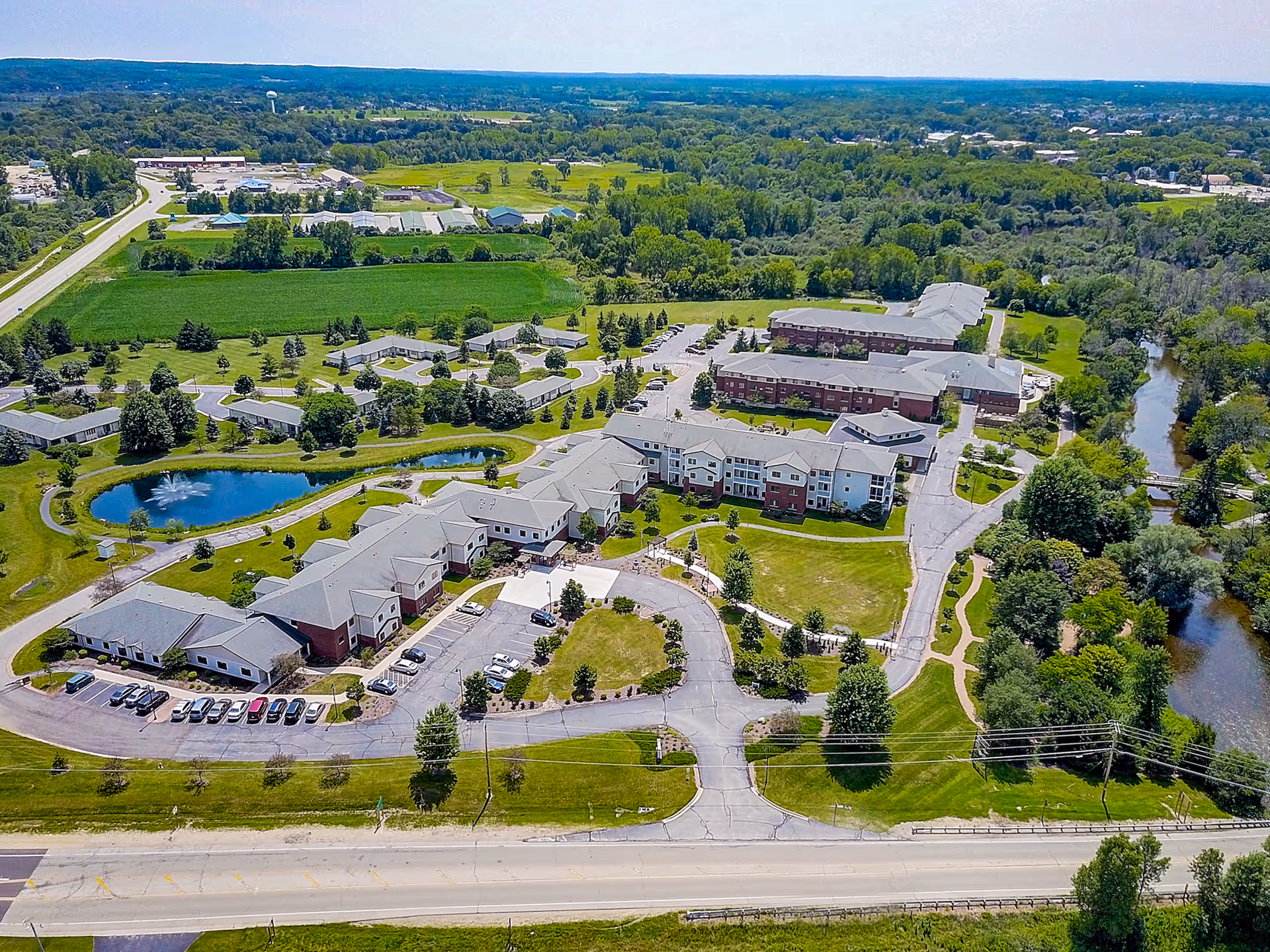 Aerial view of Three Pillars Senior Living Communities showing multiple buildings surrounded by green lawns, trees, a pond with a fountain, parking lots, and a nearby river. The facility is located near a main road with open fields and wooded areas in the background.
