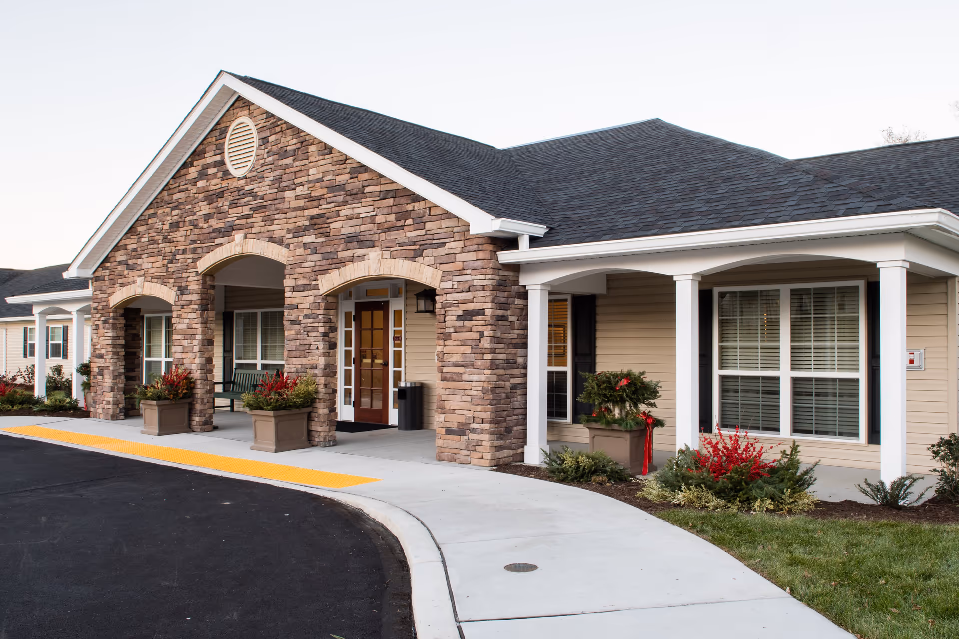 Exterior view of a senior living facility entrance with stone facade, beige siding, white columns, and a covered porch. There are potted plants with red flowers and greenery near the entrance and windows with white blinds.