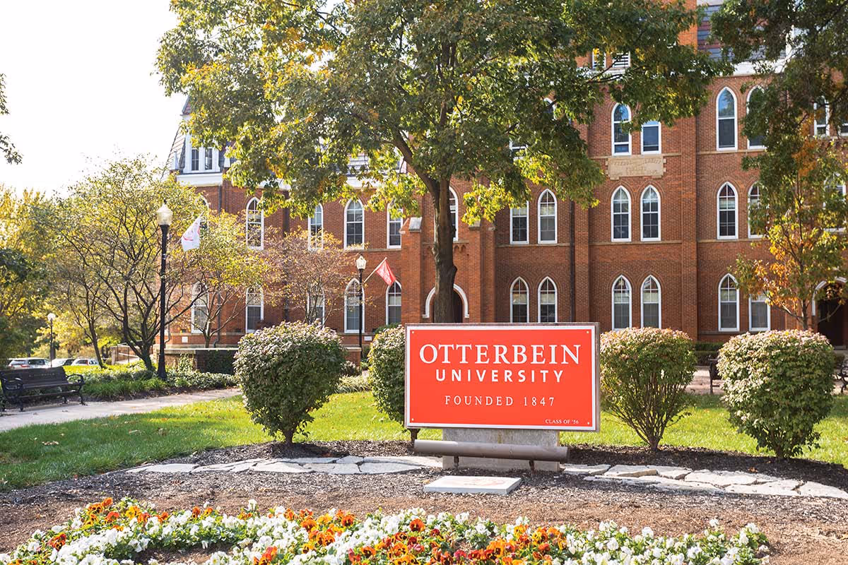 A red sign reading 'Otterbein University Founded 1847' is displayed in front of a large brick building with arched windows. The area around the sign features green grass, bushes, trees, and a flower bed with colorful flowers. There are benches and lampposts along a pathway in the background.