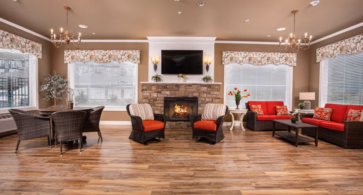 A cozy living room area in Deer Ridge Memory Care featuring a stone fireplace with a TV mounted above it, two cushioned wicker chairs with red seats in front of the fireplace, a wicker sofa and loveseat with red cushions and floral pillows, a coffee table, side tables with lamps and flowers, a round dining table with wicker chairs, large windows with floral valances, and wooden flooring.