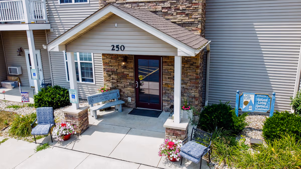 Entrance of a senior living facility with the address number 250 above the door. The entrance has a small covered porch with stone pillars, a bench, two chairs with cushions, and flower pots on either side. There is a sign on the right side indicating it is an assisted living apartment facility. The building exterior is light gray with stone accents around the door.