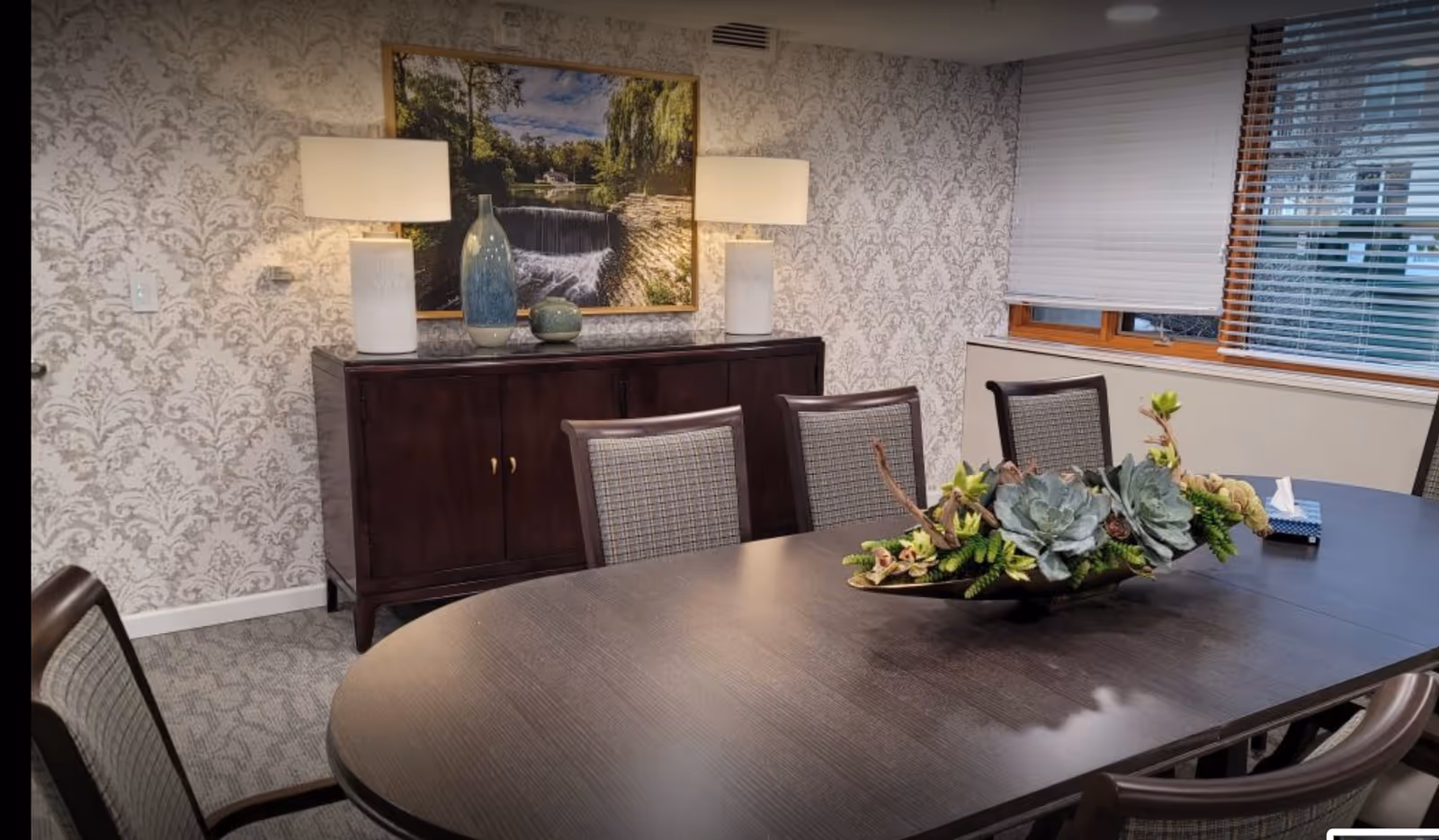 A dining room with a dark wooden oval table surrounded by six upholstered chairs. On the table is a decorative centerpiece with succulents and greenery. Against the wall is a dark wooden cabinet with two white lamps and decorative vases. Above the cabinet hangs a framed picture of a waterfall and trees. The walls have a patterned wallpaper, and there is a window with blinds partially open.