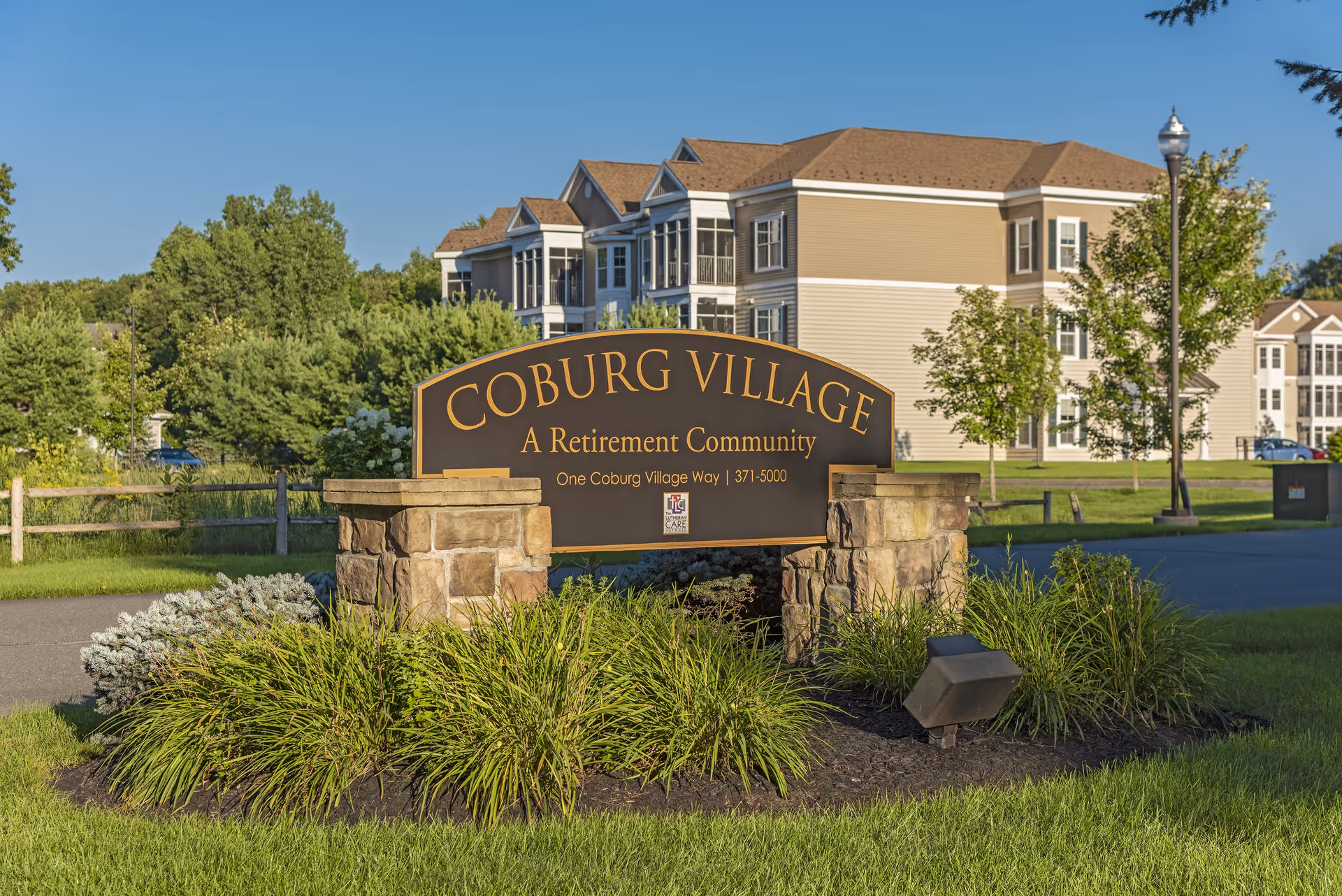 Entrance sign for Coburg Village, a retirement community, with a large building and trees in the background under a clear blue sky.