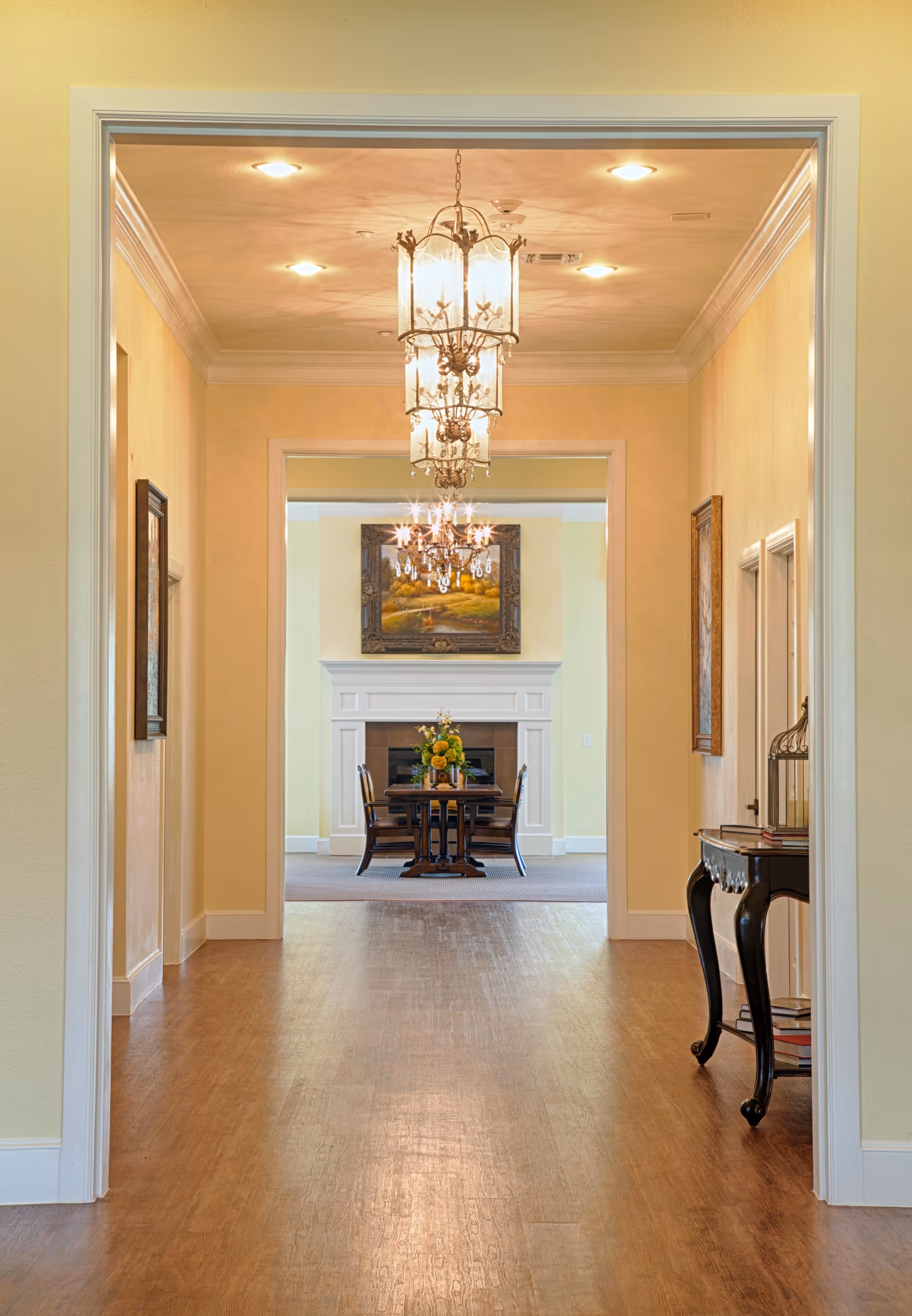 View down a hallway with wooden flooring leading to a dining area with a table and chairs in front of a white fireplace. The hallway walls are painted light yellow and adorned with framed artwork. Elegant chandeliers hang from the ceiling, and a decorative console table with books and a birdcage is positioned on the right side of the hallway.