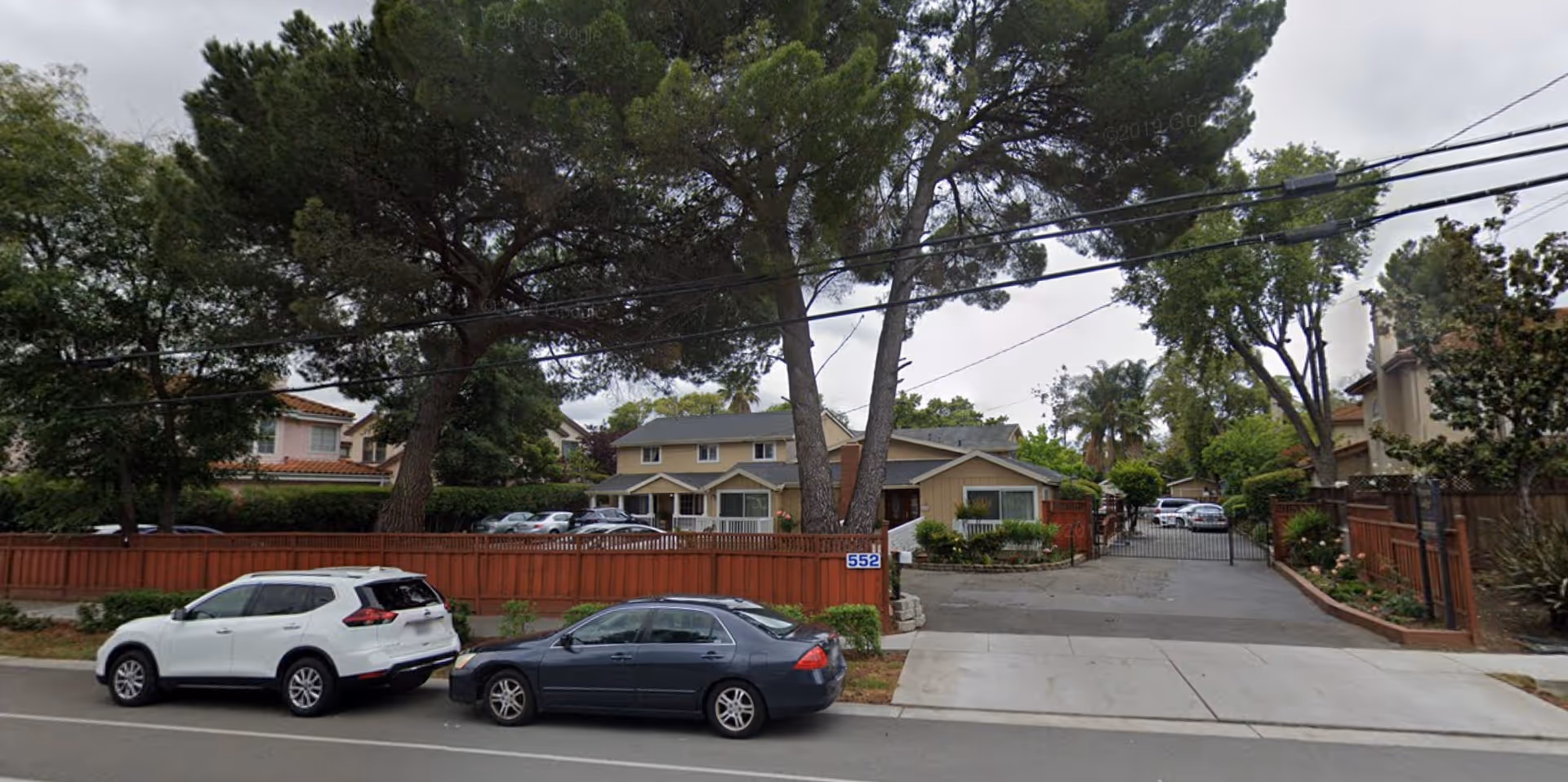 View of a residential-style building behind a wooden fence with a gated driveway, surrounded by tall trees and parked cars along the street in front.
