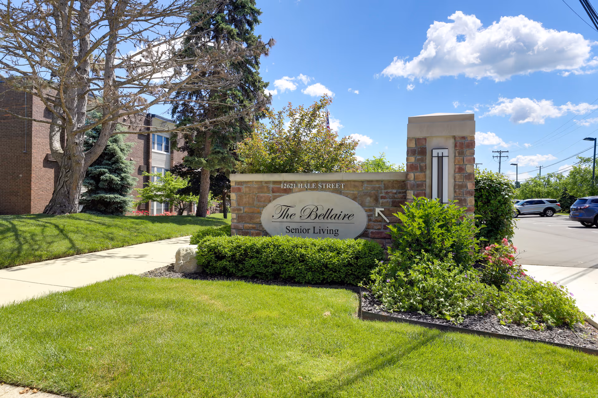 Outdoor view of The Bellaire Senior Living facility sign surrounded by green grass, bushes, and trees under a blue sky with some clouds. A sidewalk runs alongside the sign and a parking lot with cars is visible in the background.