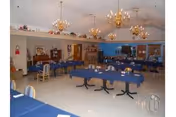 A spacious dining room with multiple rectangular tables covered in blue tablecloths, surrounded by wooden chairs. The room features several ornate chandeliers hanging from the ceiling, a piano against one wall, and various decorative items and furniture along the walls.