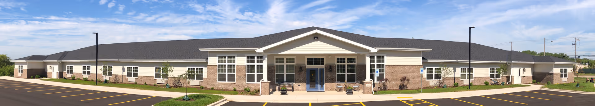 Wide panoramic view of a single-story senior living facility building with a brick and beige exterior, multiple windows, and a central entrance under a small gabled roof. The building is surrounded by a paved parking lot with marked parking spaces and a few street lamps. The sky is partly cloudy with blue patches.