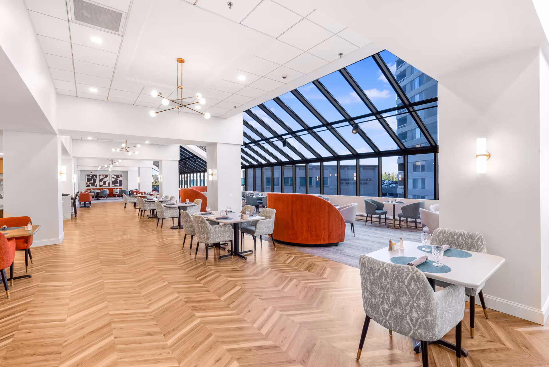 Bright and spacious dining area with modern decor featuring wooden herringbone flooring, multiple tables with gray and orange chairs, and a large glass ceiling and wall providing natural light and a view of the outside buildings.