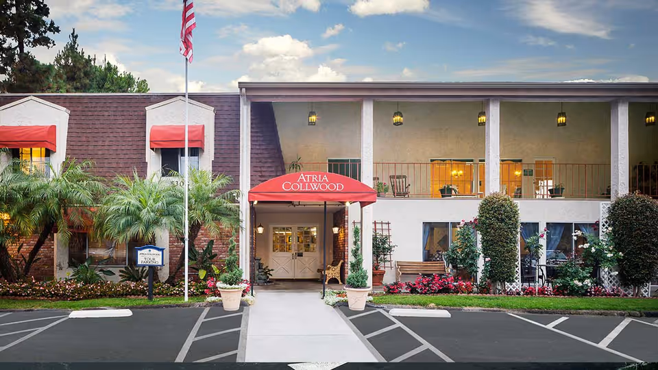 Front exterior view of Atria Collwood facility with a red awning over the entrance, an American flag on a flagpole, palm trees, and landscaped flower beds. The building has two stories with visible windows and outdoor seating on the upper balcony.