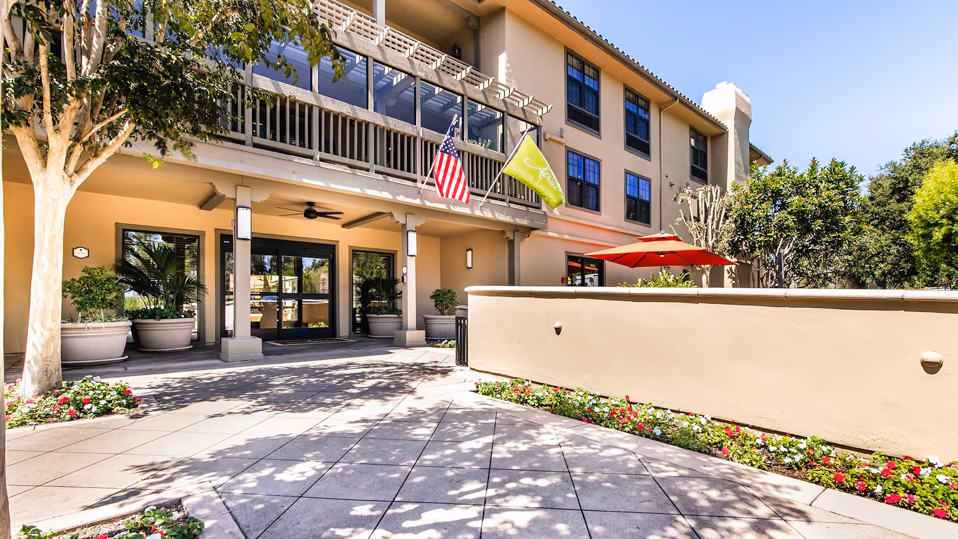 Exterior view of Atria San Juan senior living facility entrance with a beige building, large windows, two flags (American and green flag), potted plants, trees, and a tiled walkway with flower beds.