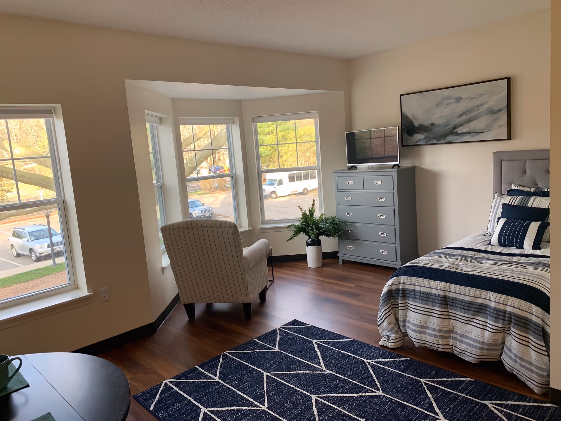 A bright and cozy bedroom with a large window area featuring three windows overlooking a parking lot. The room has a comfortable bed with striped bedding in shades of blue and white, a gray chest of drawers with a small TV on top, a potted plant beside the chest, and a cushioned armchair facing the windows. The floor is wooden with a blue and white geometric patterned rug.