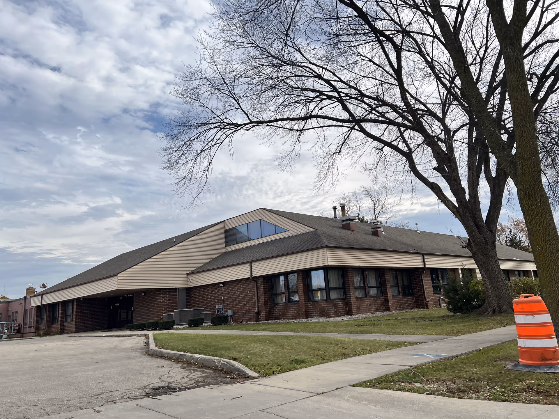 Exterior view of a single-story brick and siding building with a sloped roof, surrounded by a sidewalk and grass. A large leafless tree is visible on the right side, and an orange and white traffic barrel is near the sidewalk. The sky is partly cloudy.