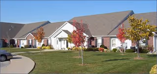 Exterior view of a single-story senior living facility building with a sloped roof, brick and white siding, small trees with autumn foliage, and a curved driveway in front.