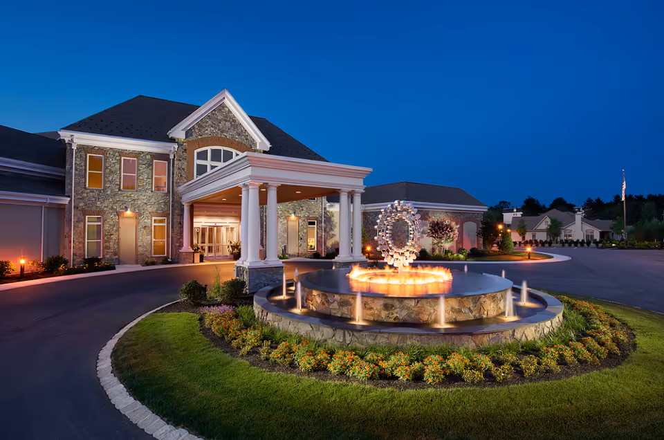 Exterior view of Manor at Willow Valley Communities at dusk, featuring a stone building with a covered entrance supported by white columns, a circular driveway, and a decorative water fountain with lights in the center surrounded by landscaping and flowers.