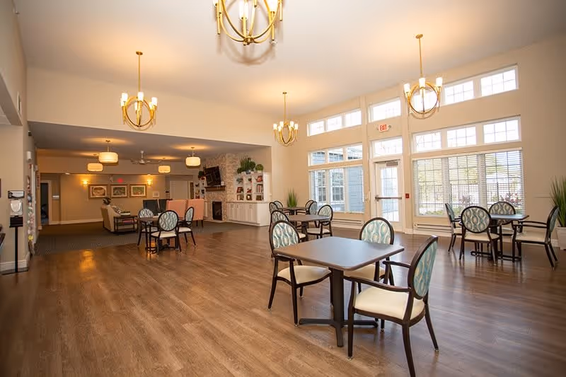 A spacious, well-lit common area in a senior living facility with several small tables and chairs arranged on a wood floor. Large windows let in natural light, and there are modern chandeliers hanging from the ceiling. In the background, there is a seating area with couches, a fireplace, and built-in shelves with decorative items.