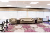 A bright common area with large windows covered by white blinds. The room features a beige sofa and two matching armchairs arranged around a small wooden table. The floor is covered with a carpet that has a circular pattern in shades of purple, beige, and brown. There are also several wooden chairs with dark frames visible on the left and right edges of the image.