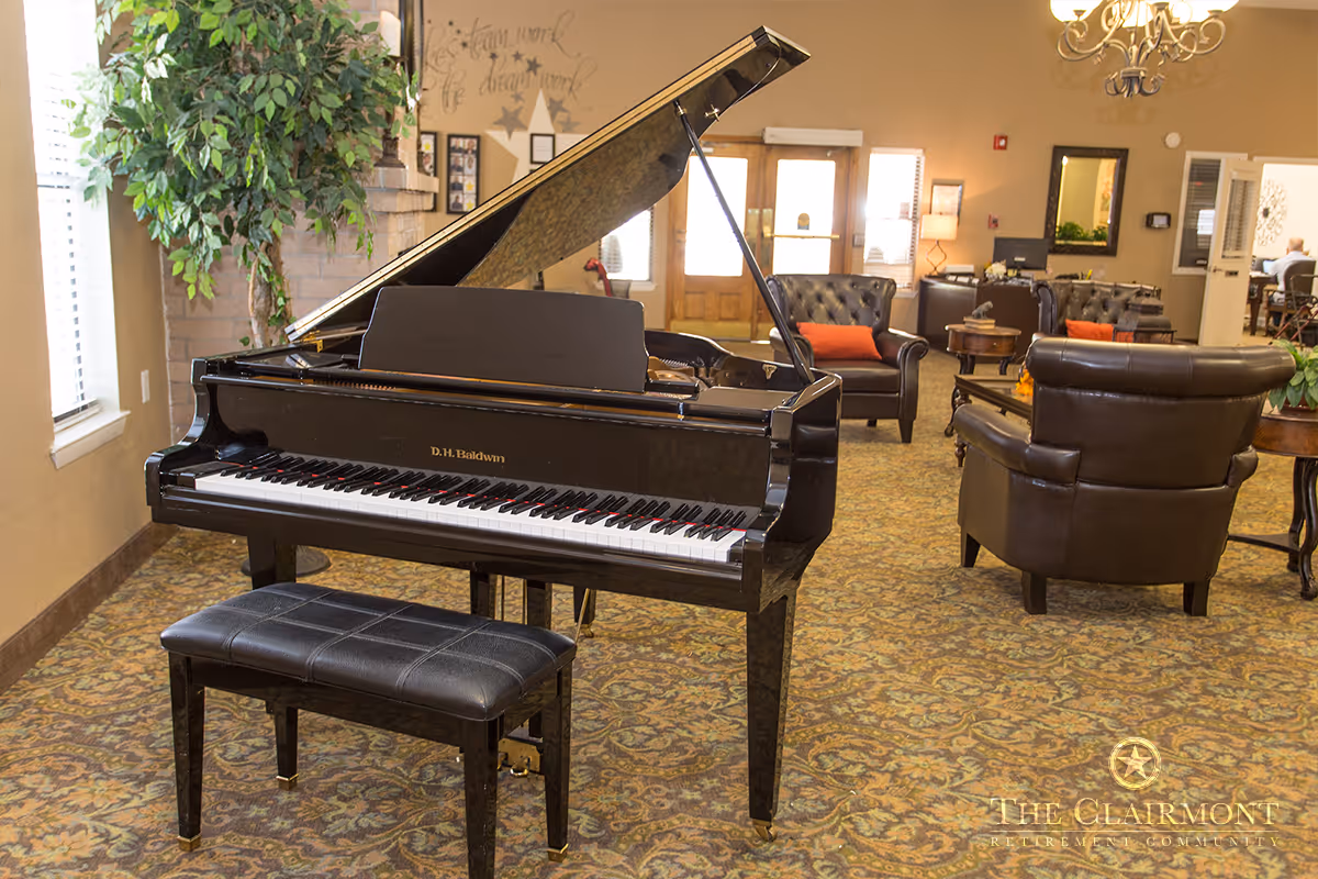A cozy common area in a retirement community featuring a black grand piano with a matching bench in the foreground. The room has patterned carpet, leather armchairs with orange cushions, a large potted plant, and a chandelier. The entrance door and windows allow natural light to brighten the space.