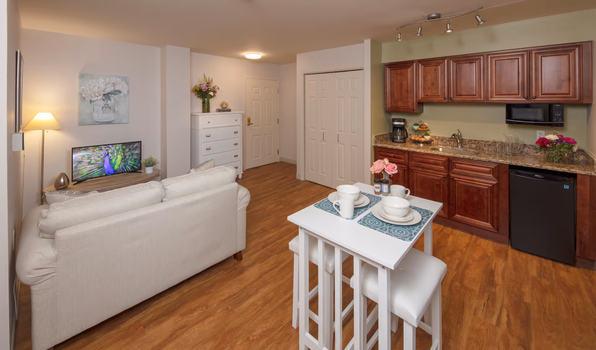 Interior view of a senior living facility apartment featuring a small living area with a beige sofa facing a TV on a wooden stand, a white chest of drawers with a vase of flowers, and a kitchenette with wooden cabinets, a granite countertop, a coffee maker, microwave, and mini fridge. In the foreground, there is a small white dining table set for two with dishes and flowers. The flooring is wood, and the walls are painted in neutral tones.