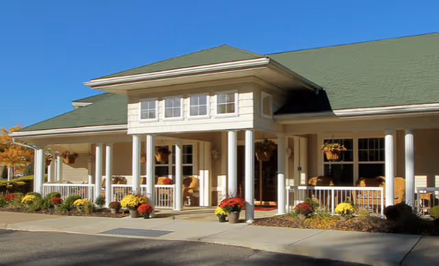 Exterior view of a single-story building with a green roof and beige siding, featuring a covered porch with white columns, hanging flower baskets, potted plants, and wicker chairs. The entrance is centered under a small gabled roof section with multiple windows.