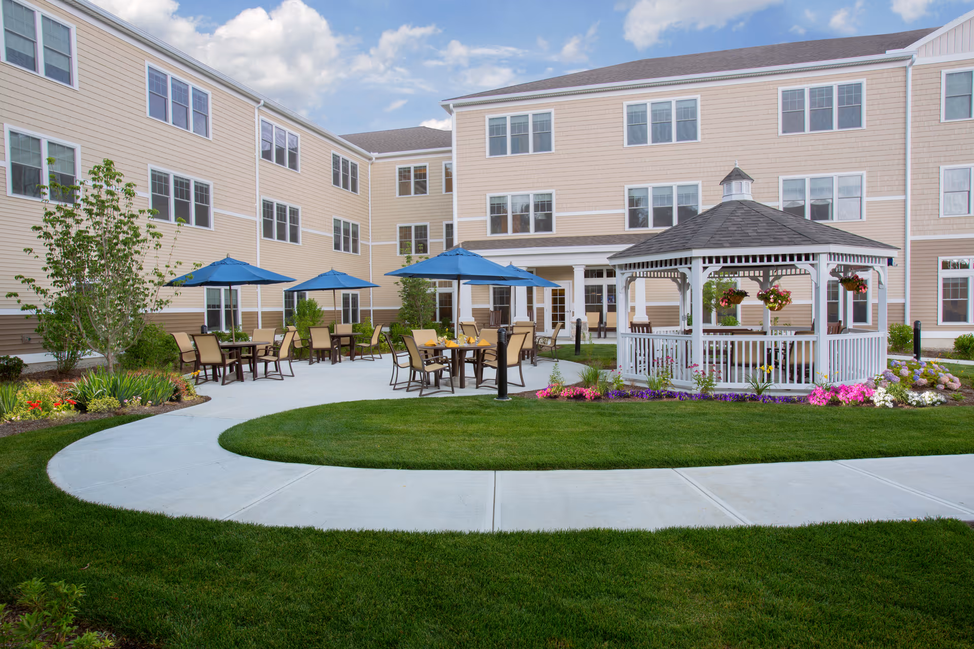 Outdoor courtyard area of a senior living facility with a white gazebo surrounded by colorful flowers, several tables with chairs and blue umbrellas, green grass, and a beige multi-story building in the background under a partly cloudy sky.