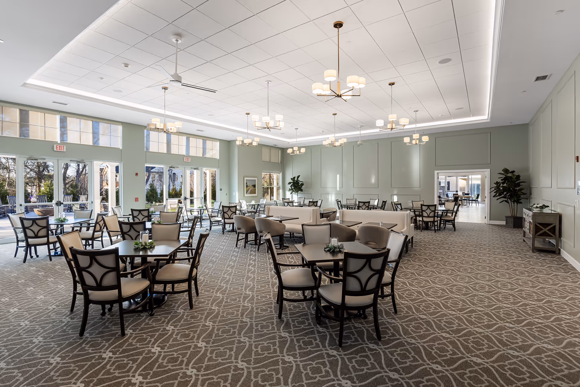 A spacious dining room with multiple tables and chairs arranged neatly. The room features large windows and glass doors allowing natural light to fill the space. The walls are painted a soft green, and the ceiling has modern light fixtures and ceiling fans. There are plants and decorative elements along the walls, and the carpet has a patterned design.