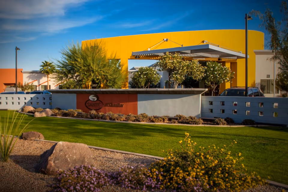 Exterior view of Santé of North Scottsdale - Skilled Nursing & Rehabilitation facility with a modern yellow and white building, landscaped green lawn, trees, bushes, and a stone sign in front displaying the facility's name.