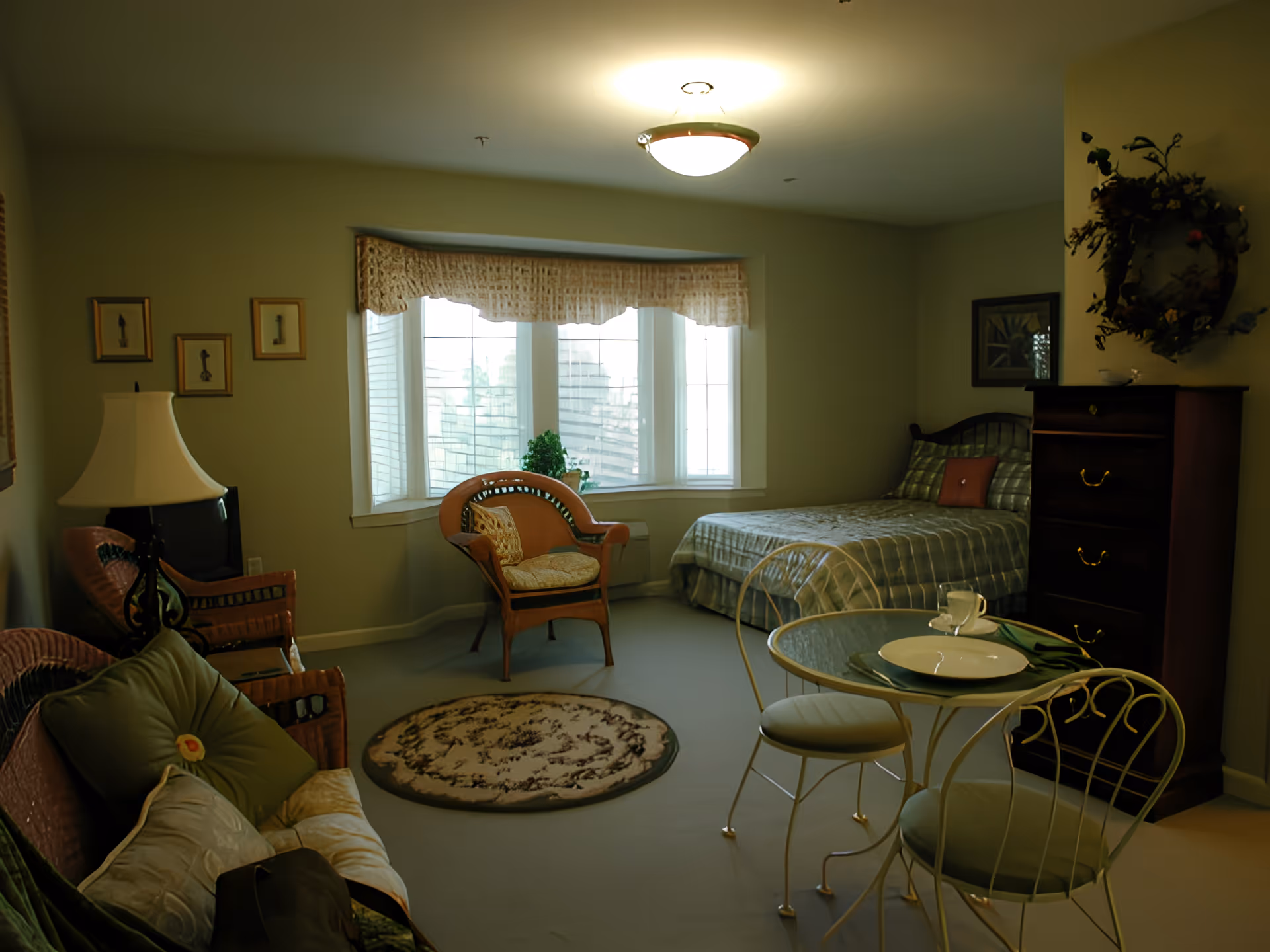 A cozy senior living room with a bed, a small round glass table set with a plate and cup, two chairs, a cushioned wicker sofa, a wicker armchair, a wooden dresser, and a window with curtains letting in natural light. The room is softly lit with a ceiling light and decorated with framed pictures and a floral wreath on the wall.