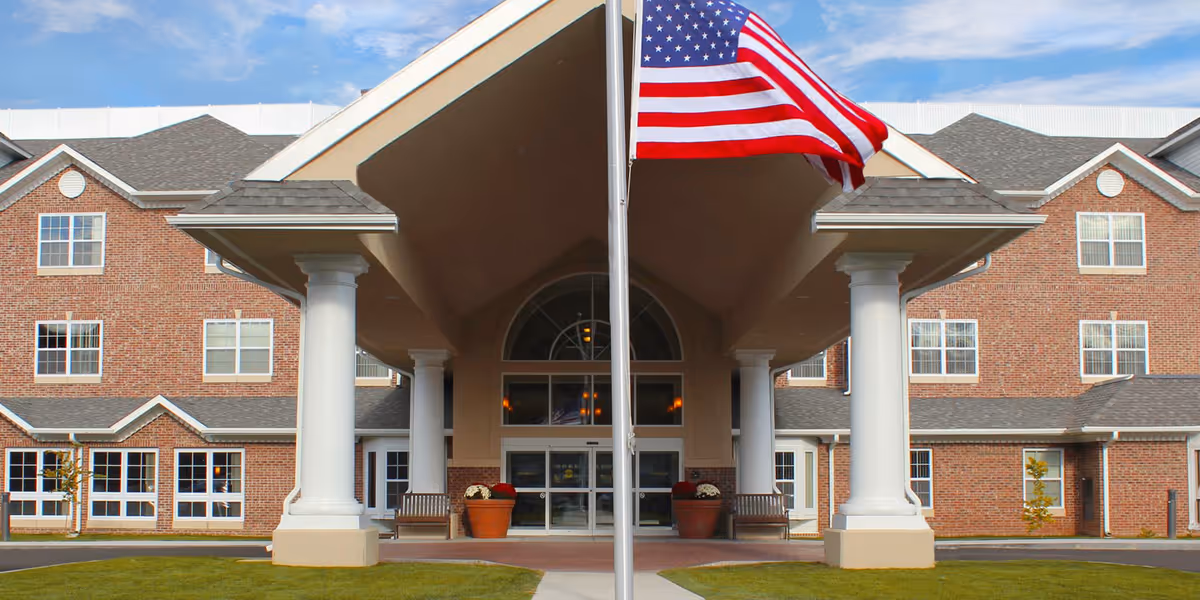 Front exterior view of a large brick retirement community building with white columns supporting a covered entrance. An American flag is prominently displayed on a flagpole in front of the entrance. The building has multiple windows and a gray shingled roof under a partly cloudy sky.