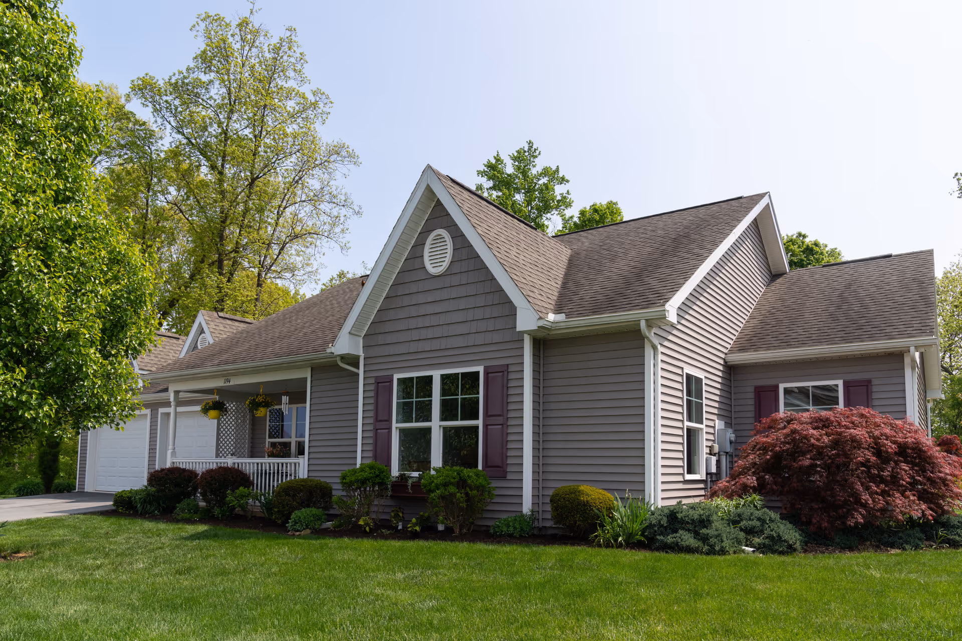 Exterior view of a single-story residential building with gray siding, white trim, and a brown shingled roof. The house has a small porch with hanging flower pots, several windows with purple shutters, and well-maintained landscaping including green bushes, a red shrub, and a lush green lawn. Trees are visible in the background under a clear sky.