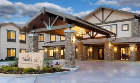 Exterior view of Paintbrush Assisted Living & Memory Care facility during dusk, showing a large covered entrance with stone pillars, warm lighting, and a sign with the facility name surrounded by landscaping.