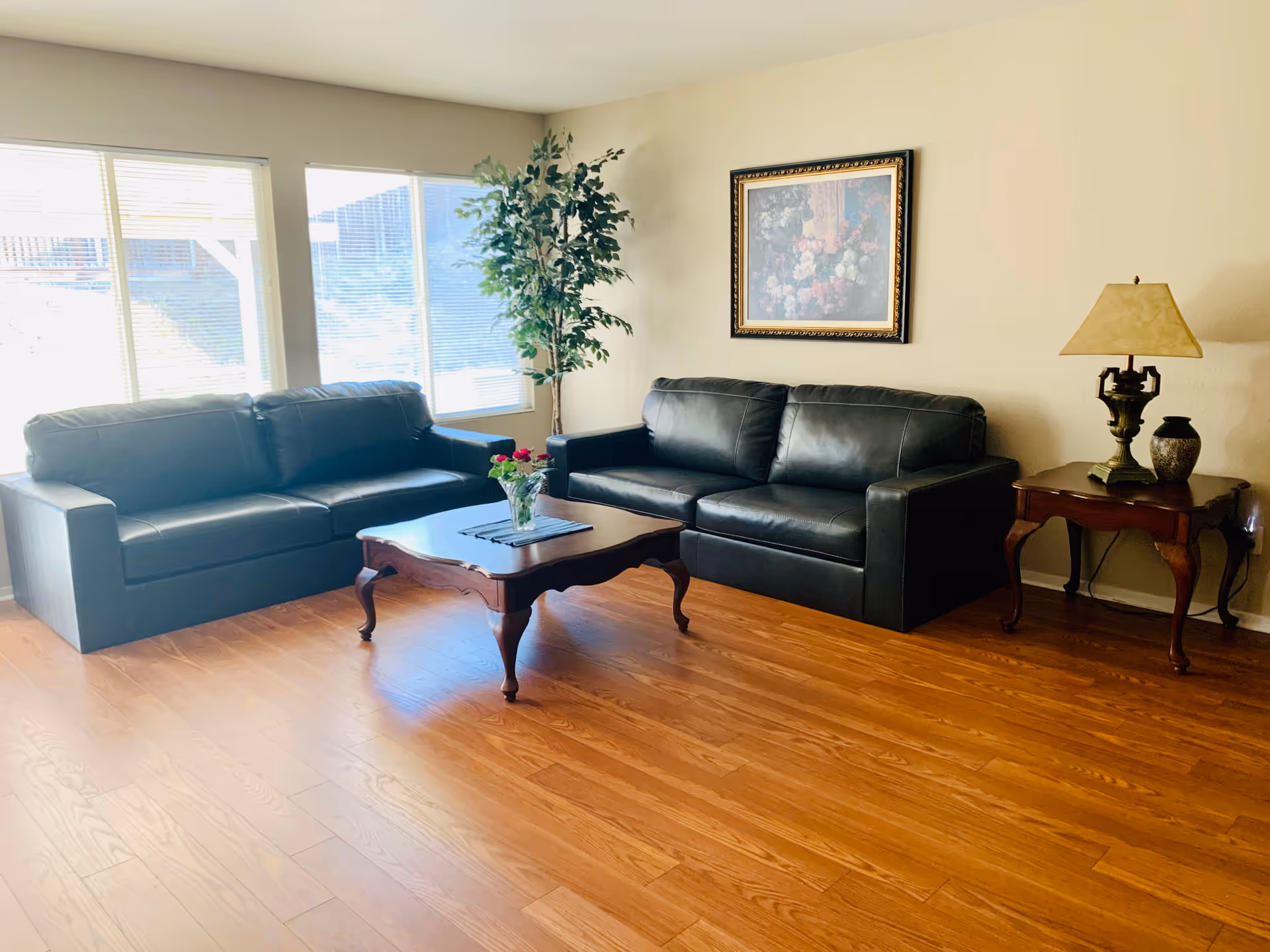 A bright living room with two black leather sofas facing each other, a wooden coffee table with a vase of flowers in the center, a side table with a lamp and decorative items, a large framed floral painting on the wall, and a tall potted plant near the windows with blinds.