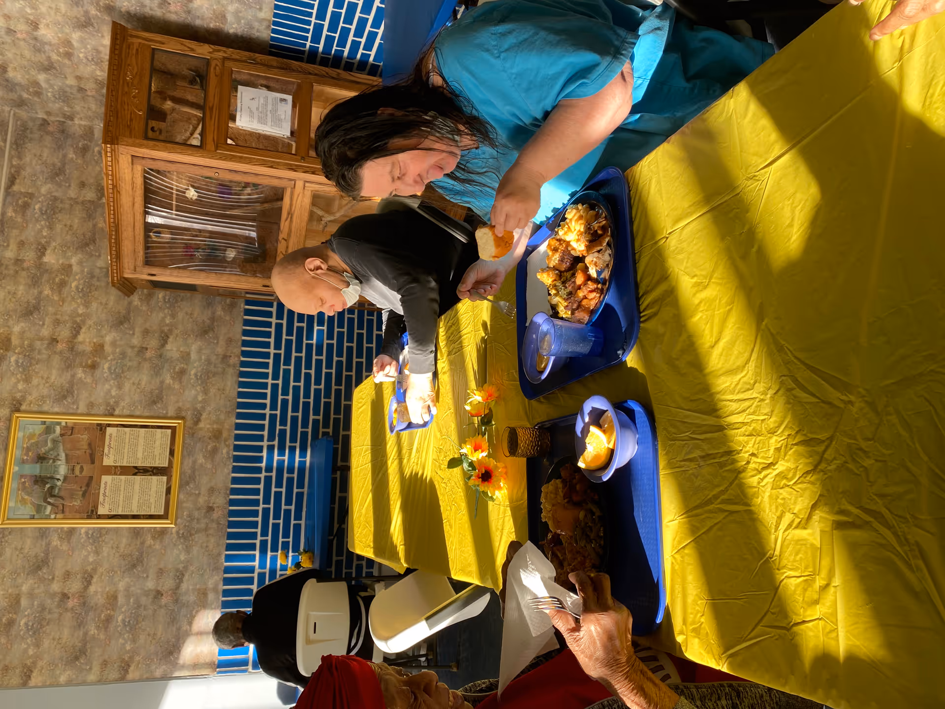 Three elderly individuals sitting at a table covered with a yellow tablecloth, eating meals served on blue trays in a dining area with blue and beige tiled walls and a wooden cabinet in the background.