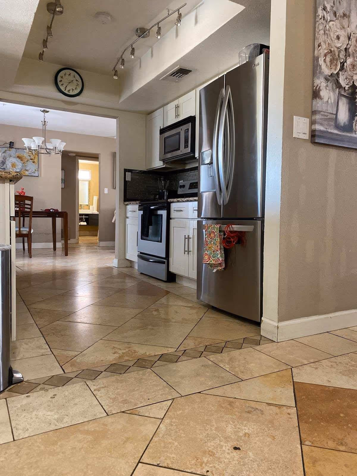 Interior view of a kitchen with stainless steel refrigerator, microwave, and stove. The kitchen has white cabinets and tiled floor with a decorative border. In the background, there is a dining area with a wooden table and chairs, a chandelier, and wall art featuring flowers. A clock is mounted on the wall above the kitchen entrance.
