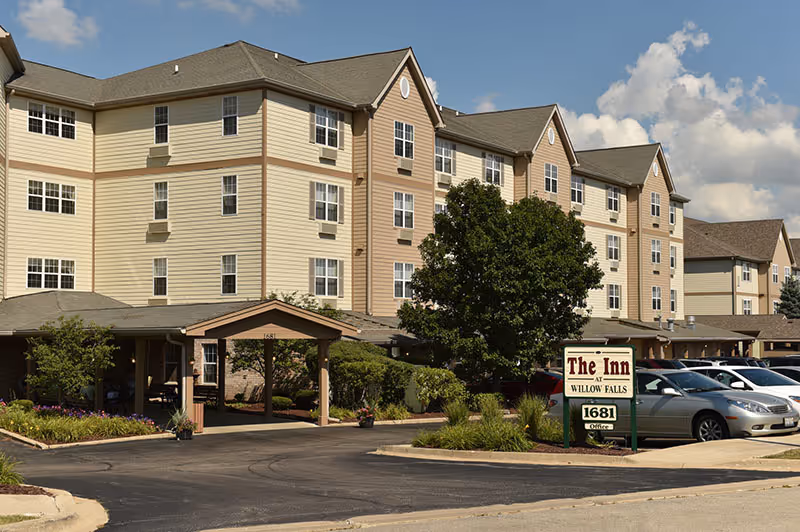Exterior view of a multi-story senior living facility named The Inn at Willow Falls, showing the building with beige and light brown siding, multiple windows, a covered entrance, landscaped greenery, and a parking lot with several cars under a partly cloudy sky.