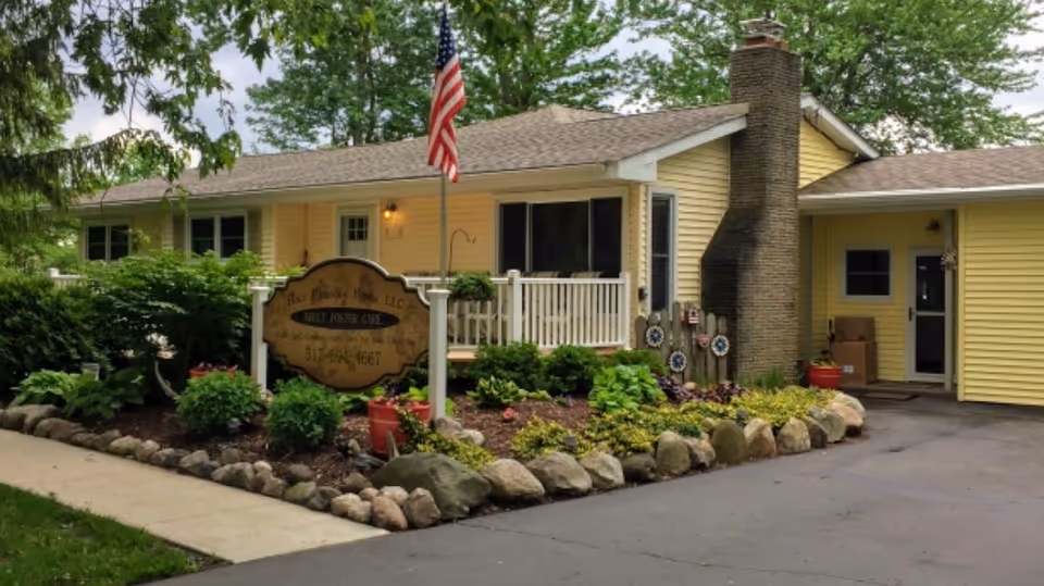 Exterior view of a single-story yellow building with a porch, surrounded by landscaped garden beds with rocks and plants. An American flag is mounted on a pole near the porch. A sign in front reads 'Holt Friendly Home, LLC Adult Foster Care' along with a phone number. The driveway and sidewalk are visible in the foreground.