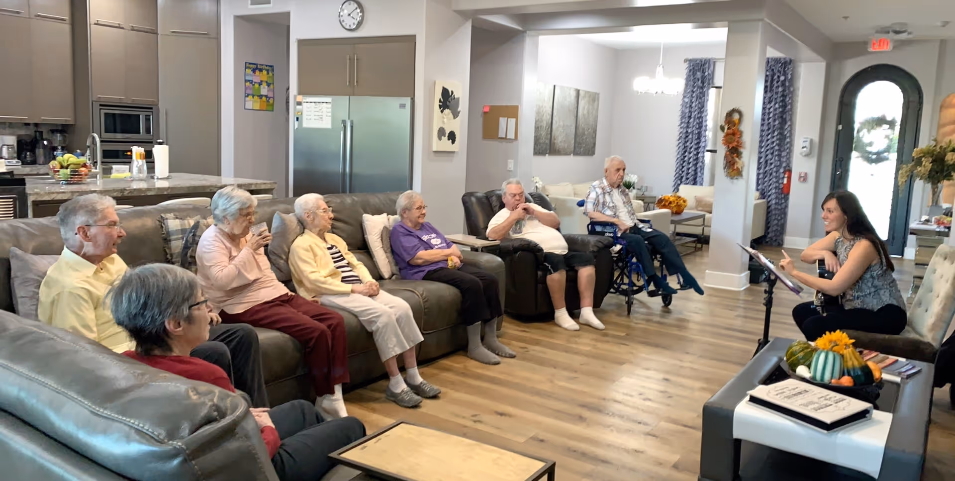 A group of elderly people seated on couches and chairs in a spacious, well-lit living room area, attentively listening to a woman who appears to be leading an activity or discussion. The room features a modern kitchen in the background, wooden flooring, and autumn-themed decorations near the entrance.
