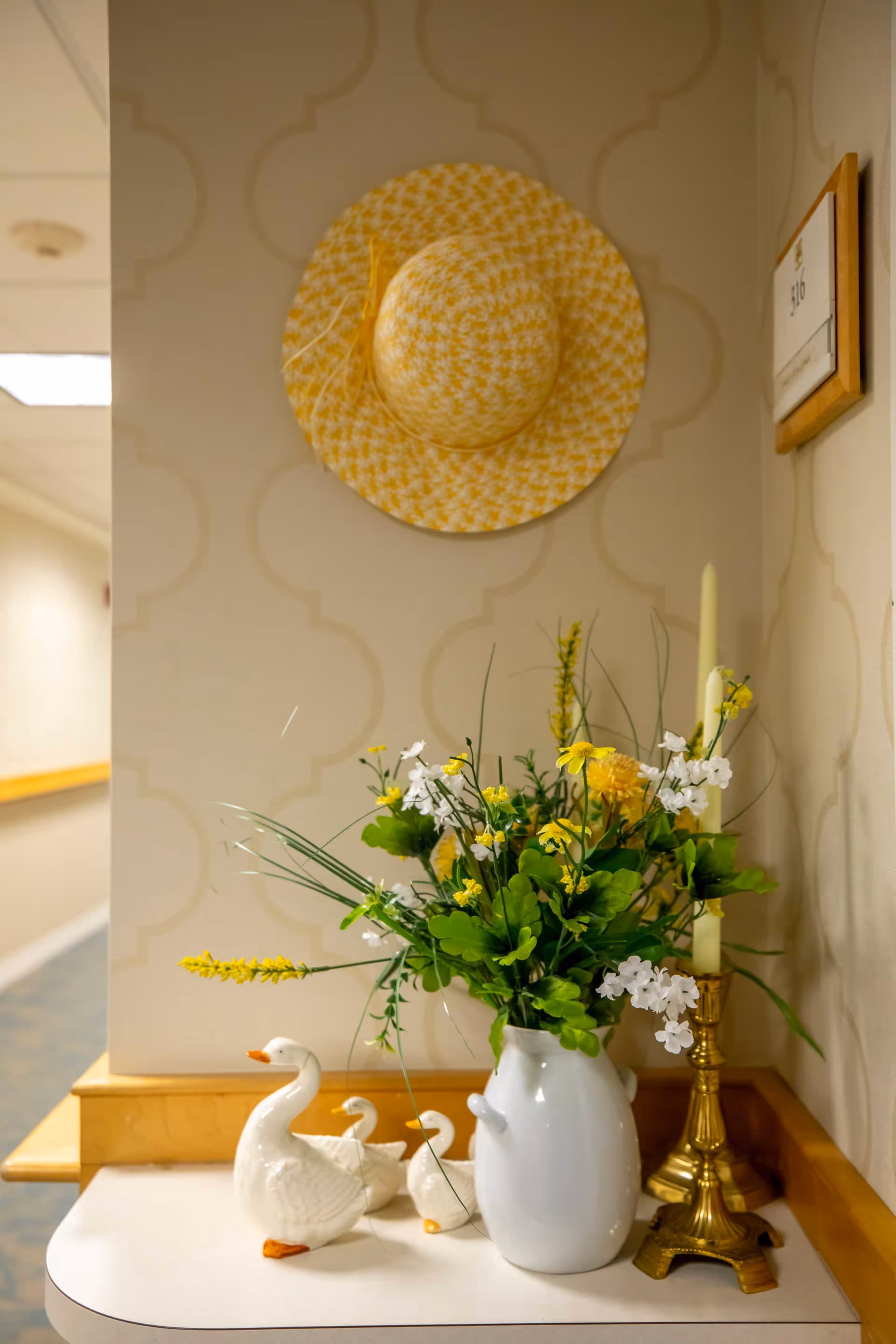 A decorative corner in a hallway featuring a yellow and white patterned hat hanging on the wall, a white vase with yellow and white flowers, three white ceramic ducks, and a brass candlestick with a white candle. The wall has a subtle geometric pattern and a room number plaque labeled 316 is visible.