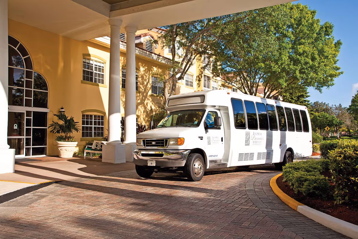A white shuttle bus parked under a covered entrance of a yellow building with large windows and white columns. There are trees and bushes around the driveway, and the bus has signage indicating it belongs to Five Star Premier Residences of Plantation.