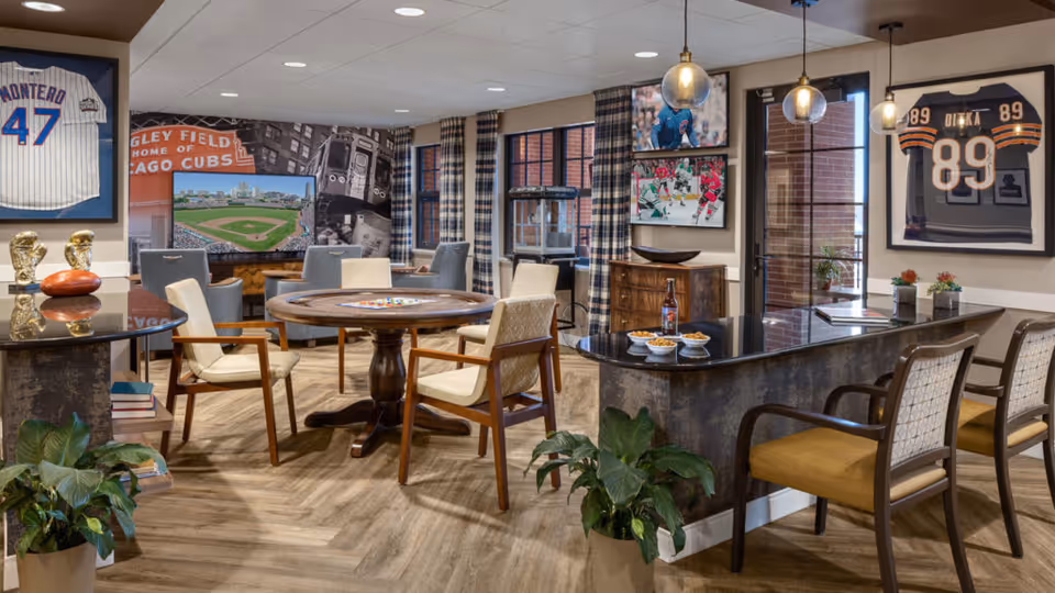 A senior living facility common area with a round wooden game table surrounded by chairs, a bar counter with stools, and sports memorabilia including framed jerseys and photos on the walls. The room has wood flooring, large windows with plaid curtains, and pendant lights hanging above the bar.