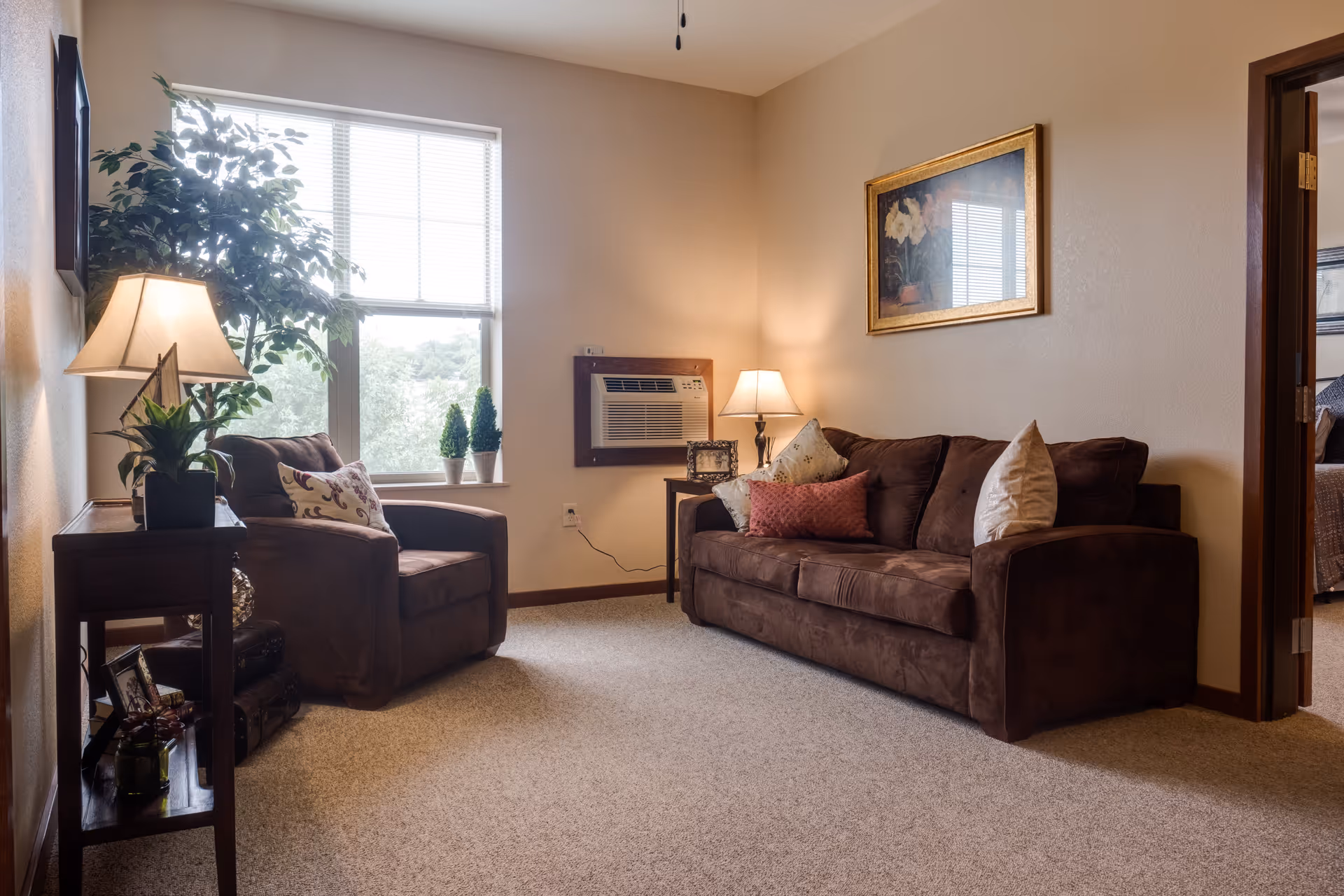A cozy living room with a brown sofa and matching armchair, both adorned with decorative pillows. There are two table lamps providing warm lighting, a large window with blinds letting in natural light, a potted plant near the window, and a framed floral painting on the wall. The room has beige walls and carpeted flooring. A doorway leads to another room with a bed visible.