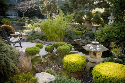 A serene garden area with neatly trimmed bushes, stone lanterns, rocks, and a black metal bench along a stone pathway surrounded by lush greenery.